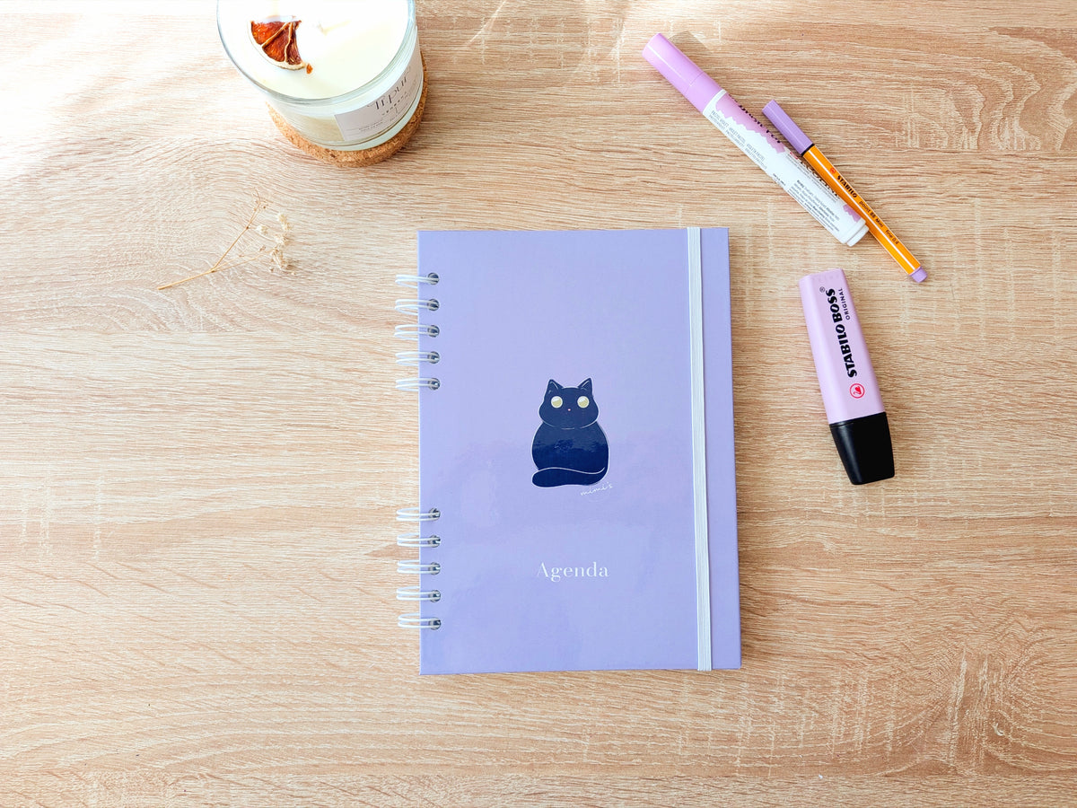 Agenda 2027 de semana vista con portada lila pastel y una ilustración de un gato negro, diseñada y encuadernada a mano.